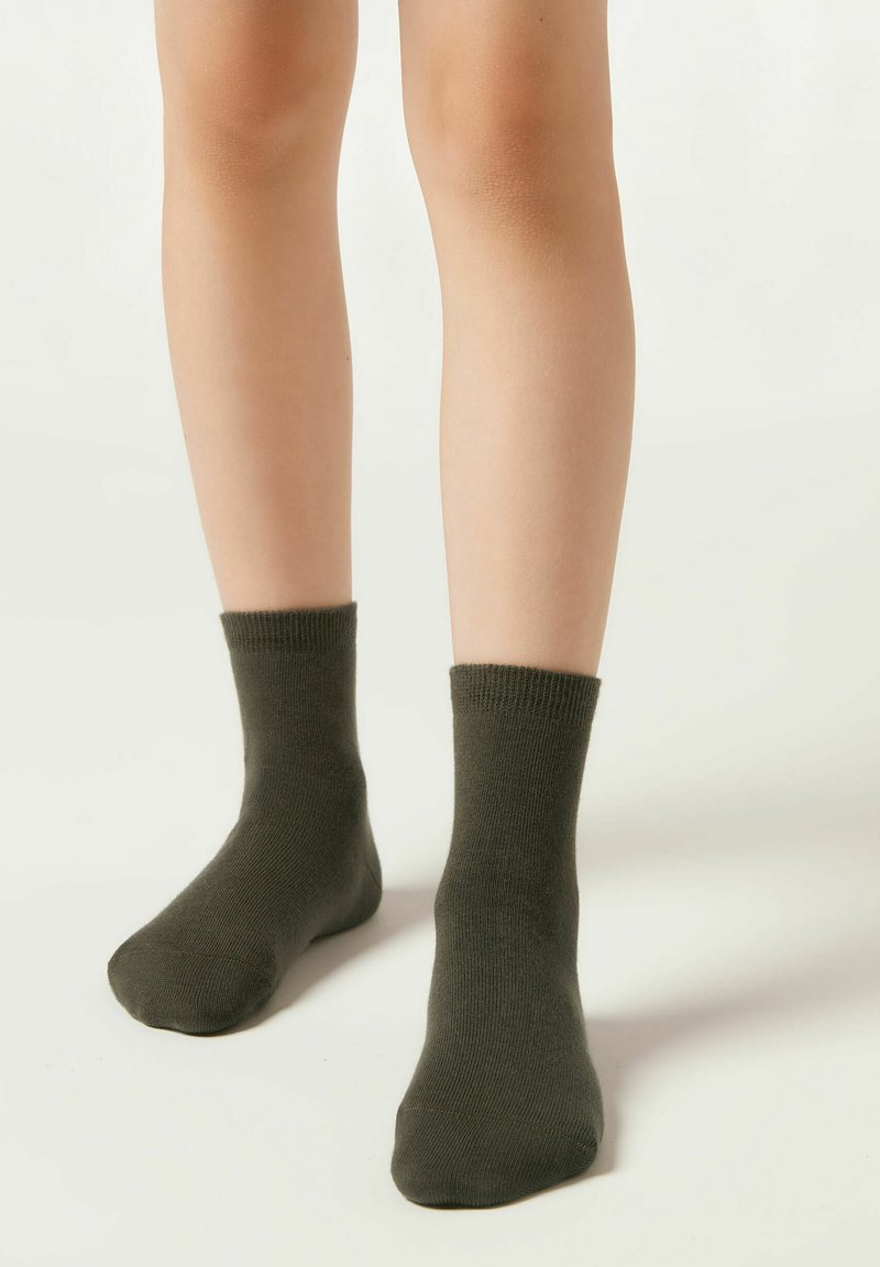 Die unteren Beine eines Kindes in dunkelgrünen Knöchelsocken, die auf einer schlichten hellen Oberfläche stehen.