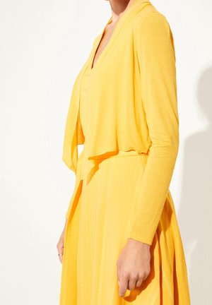 Nainen, jolla on pitkähihaisen keltainen neuletakki yllään ja sen kanssa yhteensopiva keltainen mekko, jossa on pystysuorat raidat, seisoo yksinkertaisen vaalean taustan edessä.