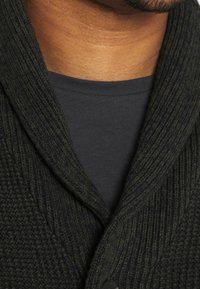 Dunkelgrüner Strickcardigan mit Schalkragen, strukturiertem Gewebe und einem schwarzen Knopf. Über einem einfarbigen dunklen Hemd getragen.