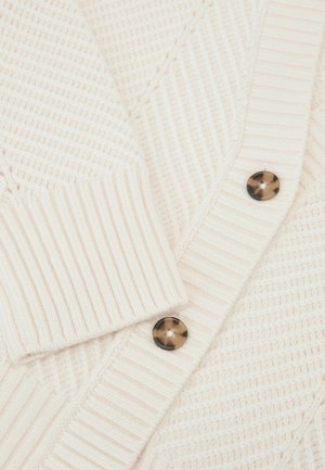 Cardigan beige clair texturé avec sections côtelées, col en V et deux boutons en écaille de tortue foncée sur le devant.