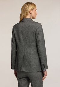 Szary żakiet z tkaniny tweedowej o dopasowanym kroju, z jednym rozcięciem z tyłu i trzema czarnymi guzikami przy każdym mankiecie. Gładka tekstura materiału i wyrafinowany fason.