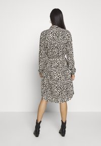 Robe portefeuille à imprimé léopard en beige et noir, avec des manches longues, une taille ceinturée et une longueur jusqu'aux genoux, accompagnée de bottines noires à la cheville.