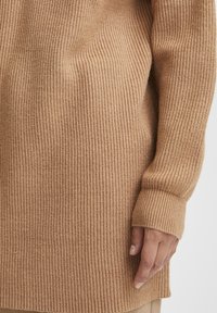 Pull en maille côtelée d'une teinte beige chaude, présentant une coupe longue et ample avec des manches longues aux poignets repliés. Texture douce et motif discret.