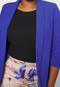 Persona que lleva un blazer azul brillante, una camiseta negra acanalada y pantalones rosa claro con estampados florales azules y morados, recortados a la altura del torso.