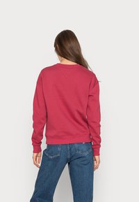 Červená mikina s kulatým výstřihem, elastickými manžetami a lemem. Vyrobena z měkké tkaniny s hladkou texturou, nošena s modrými džínami.