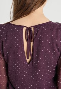 Lila Bluse mit weißen Polka-Dots, ausgestattet mit einem tiefen V-Ausschnitt am Rücken und einer Bindeschließe. Leichter, fließender Stoff mit durchsichtigen Ärmeln.