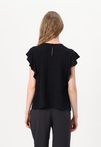 Black top with ruffled sleeves and a keyhole back detail. Made of lightweight fabric with subtle textured dots. Paired with dark, textured pants.