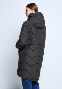 Donna che indossa un lungo cappotto imbottito nero con cappuccio, in profilo verso sinistra, su uno sfondo chiaro e uniforme.