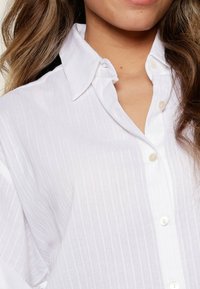 Camicia bianca con bottoni, a righe verticali chiare, con colletto classico e bottoni simili a perle, realizzata in un morbido tessuto dal tocco liscio.