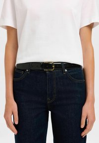 Ceinture noire avec boucle dorée portée sur un t-shirt blanc et un jean bleu foncé. La ceinture a une texture lisse et des trous ajustables.