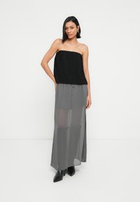 Γυναίκα που φοράει ένα μαύρο φθινοπωρινό τοπ χωρίς ράντες, συνδυασμένο με μια διάφανη γκρι μάξι φούστα και μαύρες μποτάκια αστραγάλου, σε φόντο λευκού χρώματος.