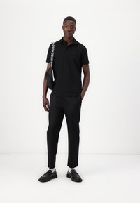 Black short-sleeve polo shirt paired with black trousers. Model holds a black bag with a white logo strap and wears black shoes. Simple design.