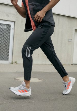 Osoba biegająca na zewnątrz, ubrana w czarne spodnie dresowe z odblaskowymi paskami oraz szare buty sportowe z jasnopomarańczowymi akcentami.