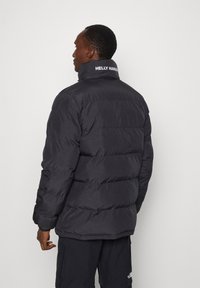 Zwarte gewatteerde jacket met hoge kraag en contrasterend logo op de rug. De stof lijkt glad met horizontale doorgestikte delen.