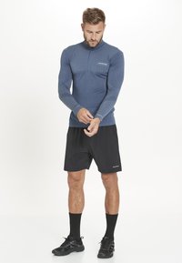 Langærmet atletisk top i mørkeblue, med en halv-zipped krave og strukturerede ærmer, parret med sorte shorts og sorte atletiske sko.