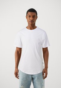 Hvit T-skjorte med korte ermer og rund hals, laget av bomull. Modellen har på seg slitte lyseblå jeans med synlige rifter.