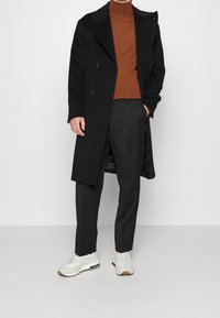 Mann mit schwarzem langen Mantel, rötlichem Rollkragenpullover, schwarzen Hosen und weißen Sneakers, der vor einem schlichten weißen Hintergrund steht.