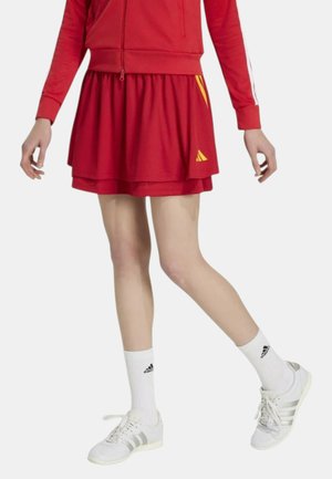 Persona con una gonna atletica rossa con dettagli gialli, calzini bianchi Adidas, scarpe da ginnastica bianche Adidas e una giacca rossa con zip.