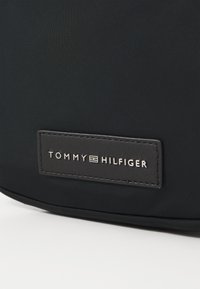Svart stoffryggsekk med teksturert overflate, med et rektangulært lærpatch med den sølvfargede "TOMMY HILFIGER"-logoen preget.