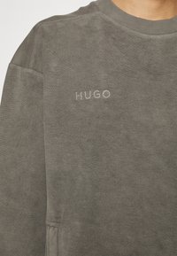 Sweat-shirt gris à col rond en tissu doux, avec un logo "HUGO" brodé. Coutures visibles et texture détaillée.