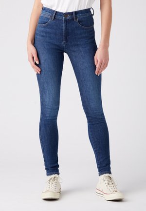 Jeans ajustados de denim azul oscuro, con una cintura alta, diseño de cinco bolsillos y detalles de costura sutiles, combinados con zapatillas blancas.