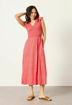 Frau trägt ein ärmelloses rotes gemustertes Midikleid mit Taschen und braunen geflochtenen Sandalen, posiert mit einer Hand hinter dem Kopf.