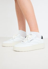 Weiße Leder Sneakers mit einer dicken cremefarbenen Sohle, perforiertem Obermaterial und schwarzen Akzenten an der Ferse. Tragen mit hellgrauen Socken.
