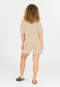 Beige texturerad kortärmad topp matchad med matchande shorts. Modell som bär bruna slip-on sandaler, sett bakifrån.