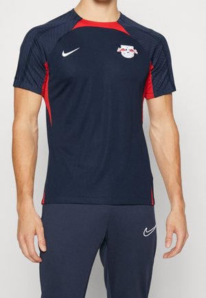 Mężczyzna w granatowej i czerwonej sportowej koszulce z krótkim rękawem oraz granatowych spodniach z białymi logo Nike, zwrócony twarzą do przodu na jednolitym tle.