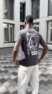 Pilkos spalvos marškiniai trumpomis rankovėmis su baltais užrašais ir dizainu ant nugaros, derinami su šviesiomis, laisvai krentančiomis kelnėmis. Plokščias modelis išdėstytas ant žemės.