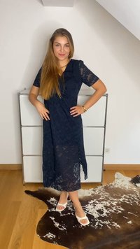 Navy lace wrap dress with short sleeves, knee-length, featuring a side slit. Paired with white flat open-toe sandals on a patterned rug.