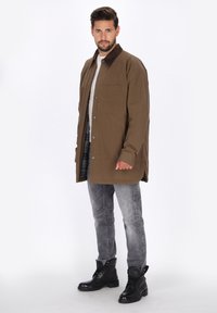 Braune button-up Jacke mit einem dunklen Kragen, zwei vorderen Taschen und einer lockeren Passform, kombiniert mit grauen Jeans und schwarzen Schnürstiefeln.