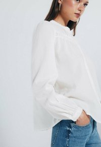 Weiße Bluse mit langen Ärmeln, gerafften Schultern, Knopfleiste und Spitzenakzenten. Kombiniert mit engen blauen Jeans mit Nietenverzierungen.