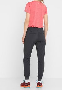 Žena v červeném tričku s krátkým rukávem, černých kalhotách Columbia, bílých ponožkách a červených běžeckých botách na trail, stojí zády k jednoduchému bílému pozadí.