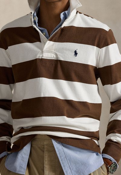 Uomo che indossa una maglia da rugby a righe larghe marroni e bianche sopra una camicia azzurra con bottoni, abbinata a pantaloni kaki e un orologio da polso.