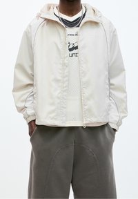 En person som bär en krämfärgad zip-up jacka över en vit nättröja med svart text och grå byxor med geometrisk sömnad, med händerna i fickorna.