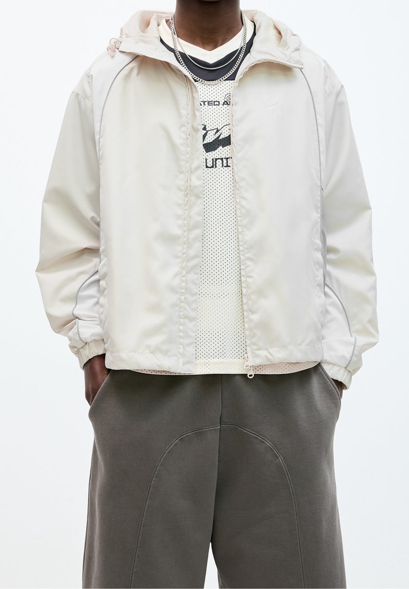 En person som bär en krämfärgad zip-up jacka över en vit nättröja med svart text och grå byxor med geometrisk sömnad, med händerna i fickorna.