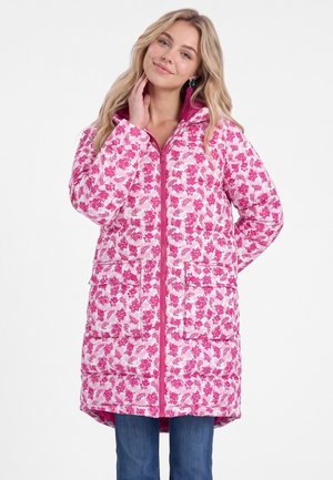 Junge Frau trägt eine lange rosa-weiße gesteppte Jacke mit Blumenmuster, großen Fronttaschen und blauen Jeans, posiert mit der Hand in der Nähe des Halses.