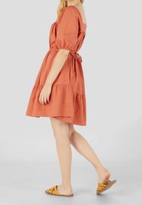 Robe en coton couleur rouille-orange avec des manches courtes bouffantes, une jupe à étages et un détail noué à la taille. Associée à des sandales plates marron avec des lanières jaunes.