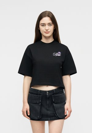 Νεαρή γυναίκα με μακριά καστανά μαλλιά, φορώντας μαύρο κοντό T-shirt με floral λεπτομέρειες στο κείμενο και μαύρη μίνι φούστα με μεγάλες μπροστινές τσέπες.