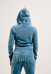 Svileni trenirku u svijetloplavoj boji, koja uključuje kapuljaču i zip zatvaranje na gornjem dijelu te odgovarajuće hlače. Ukrašena je kristalićima s natpisom "Juicy" na leđima.