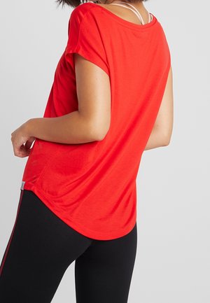 Nainen, joka pukeutuneena kirkkaanpunaiseen lyhythihaiseen toppiin ja mustiin leggingsseihin, joissa on punainen raita, esittäen selkänsä ja sivuprofiilinsa yksiväristä taustaa vasten.