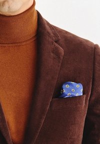 Brązowa marynarka z korduroju na pomarańczowym golfie, z niebieską poszetką w białe kwiaty.