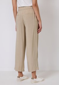Beige weite Hose mit elastischem Bund und zwei Gesäßtaschen, aus leichtem Stoff, kombiniert mit weißen Sneakern.