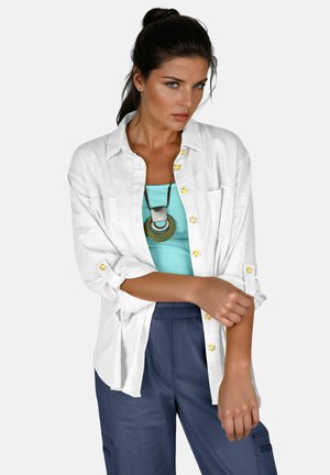 Weißes Leinenhemd mit gelben Knöpfen, darüber ein türkisfarbenes Tanktop, kombiniert mit marineblauen Cargo-Hosen. Auffälliges rundes Halskettendetail.