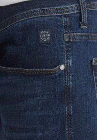 Mörkblå denimjeans med kontrasterande orange sömmar, en liten ficka och en fyrkantig tygetikett med texten "DENIM BLEND" på baksidan.