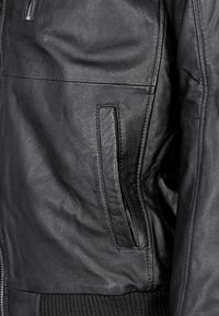 Schwarze Lederjacke mit einer glatten Textur, ausgestattet mit einer Reißverschlusstasche und geripptem Saum, die elegante Linien und ein minimalistisches Design zeigt.