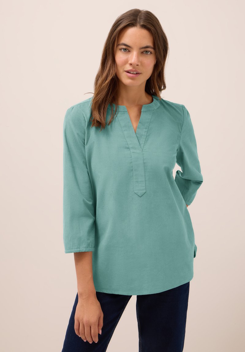 Donna con lunghi capelli castani che indossa una blusa a maniche lunghe color teal e pantaloni scuri, in piedi su uno sfondo beige semplice.