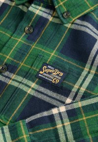 Zeleno-modrá kostkovaná flanelová košile s žlutými pruhy, se tmavými knoflíky a námořní modrou etiketou s nápisem "SuperDry". Měkká textura.