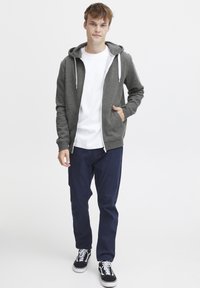 Grijze zip-up hoodie met een trekkoordcapuchon, witte voering, gedragen over een wit t-shirt. Gepaard met marineblauwe broek en zwarte sneakers.